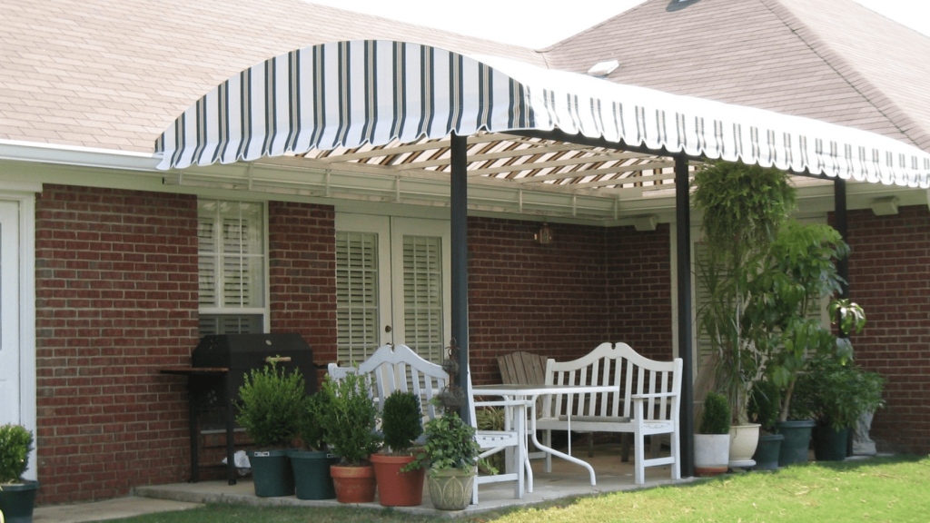 Patio with white outdoor furniture and potted plants under a striped tunnel awning attached to a brick house