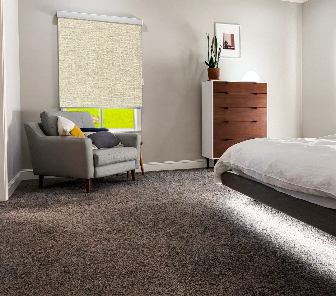 Cozy bedroom featuring a gray armchair, wooden dresser, window with a light beige roller shade, and a bed with white linens. Bright and inviting.