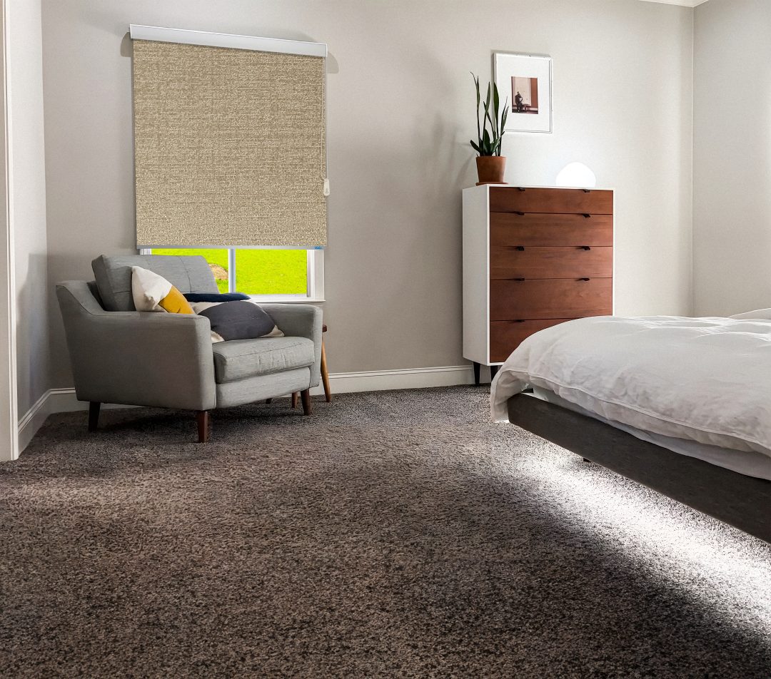 Cozy bedroom featuring a gray armchair, wooden dresser, window with a beige textured roller shade, and a bed with white linens. Bright and inviting.