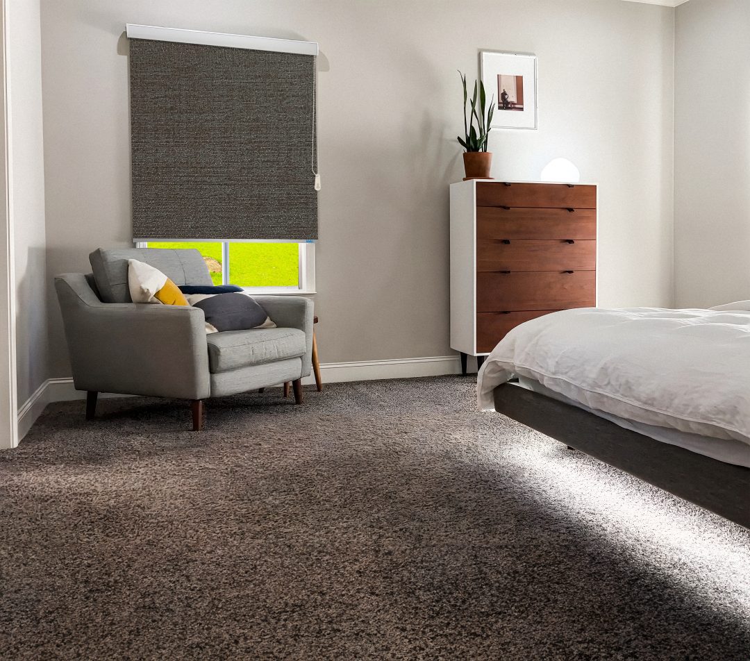 Cozy bedroom featuring a gray armchair, wooden dresser, window with a dark roller shade, and a bed with white linens. Bright and inviting.