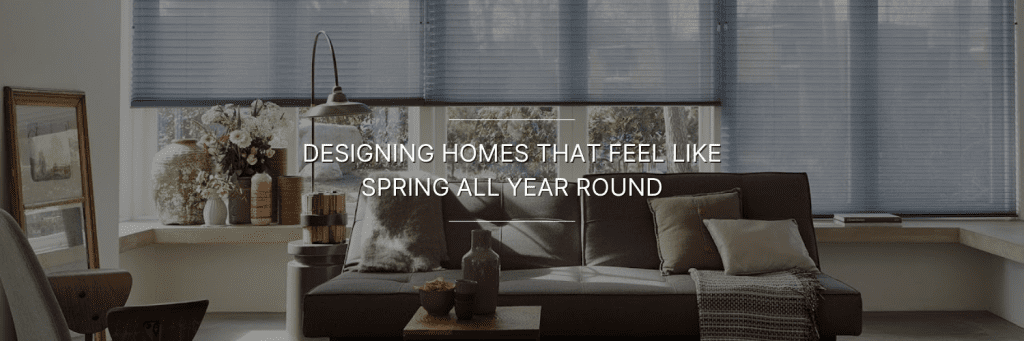 Cozy living room with a brown sofa, cushions, a vase on a wooden table, and large windows with blinds. Text reads “Designing homes that feel like spring all year round.”