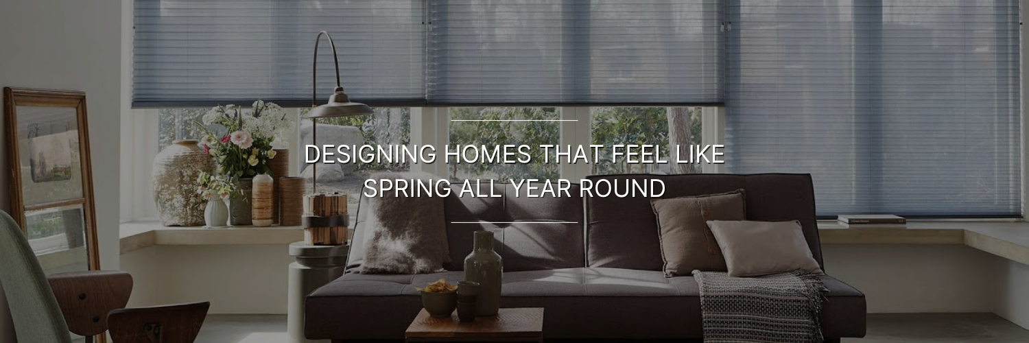 Cozy living room with a brown sofa, cushions, a vase on a wooden table, and large windows with blinds. Text reads “Designing homes that feel like spring all year round.”