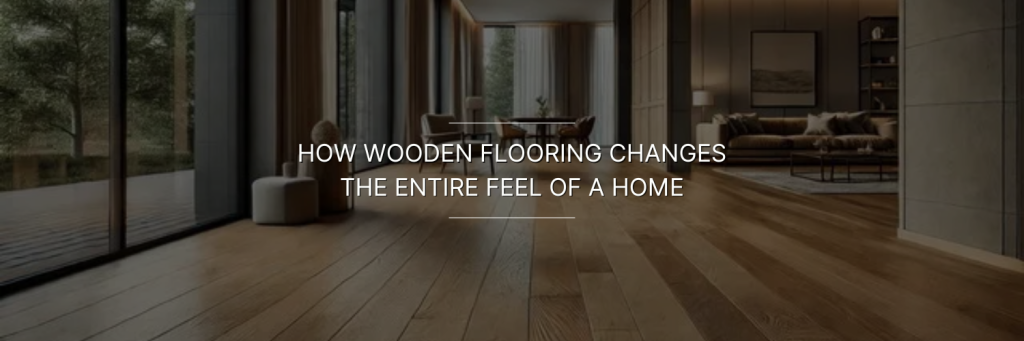 Wooden-floored living room with large windows overlooking a forest. Cozy furniture and soft lighting create a warm, inviting atmosphere. Text overlay reads: "How wooden flooring changes the entire feel of a home.